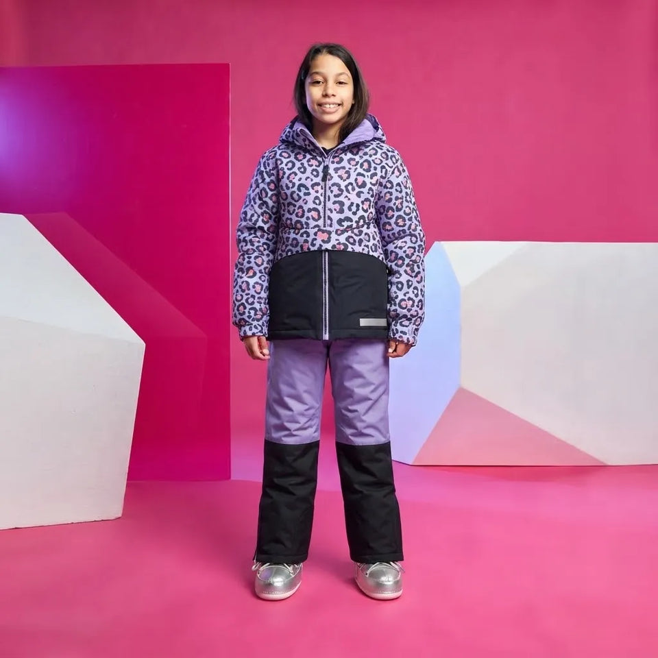A young girl stands in a studio wearing a children's snow jacket and snow pants. The jacket has a purple and black leopard print pattern on the upper portion and a solid black lower panel.