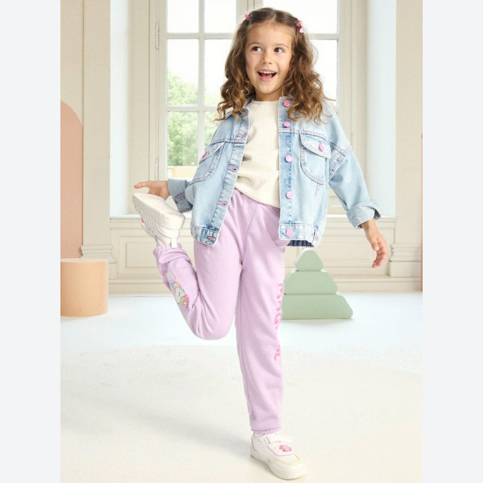 A young girl with curly brown hair stands in a bright room, stretching her leg while wearing a light blue denim jacket, cream t-shirt, lavender sweatpants, and white sneakers.