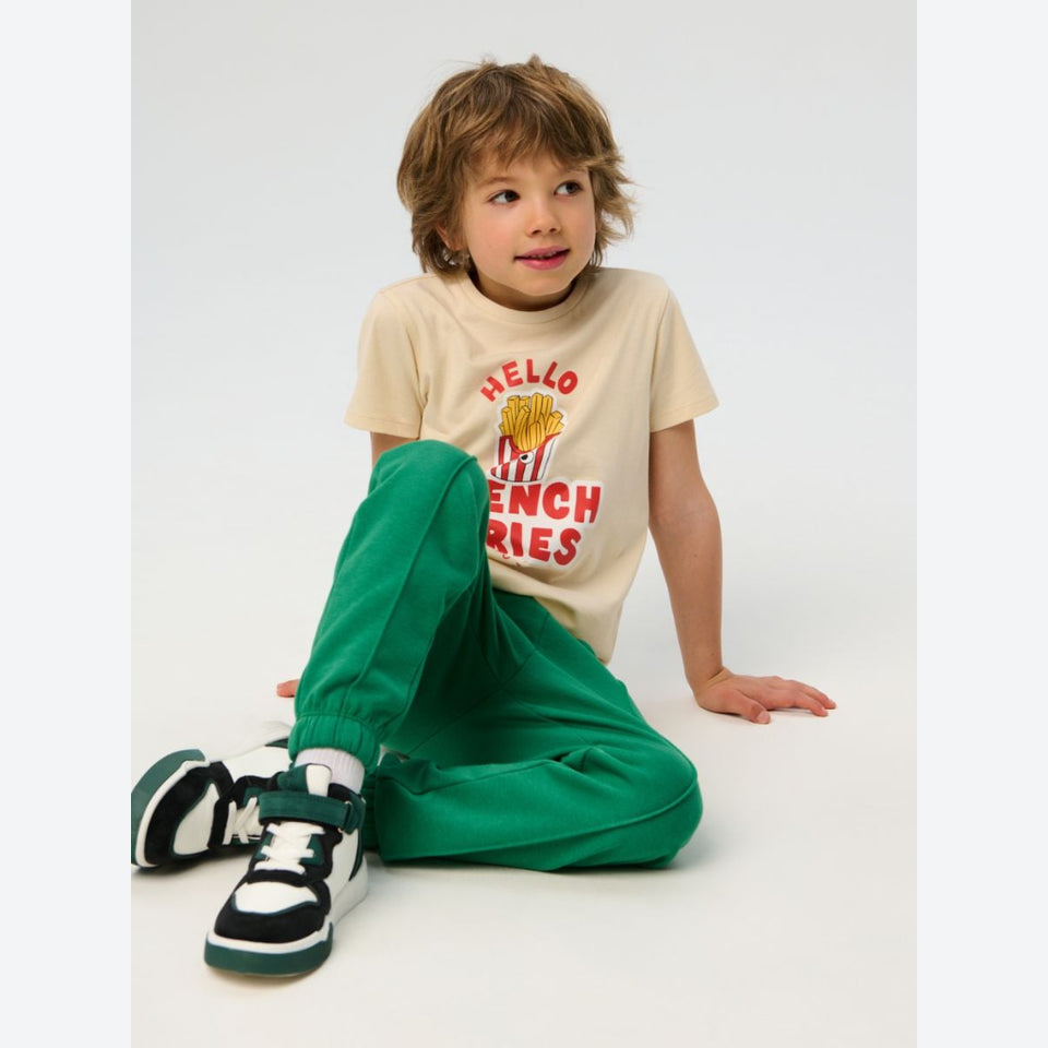 A young boy sits on a white studio floor wearing a beige graphic t-shirt with a red and yellow French fries print, bright green jogger pants, and black and white sneakers.