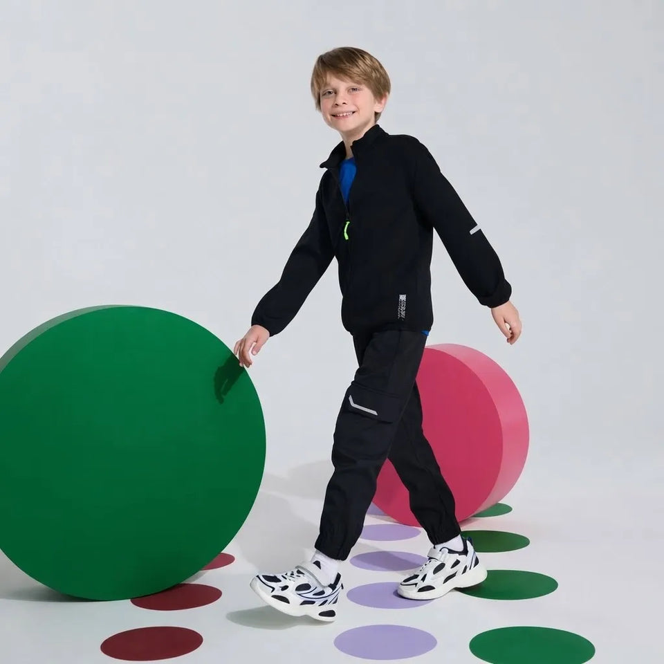 A young boy wearing a black zip-up jacket and black pants walks across a studio floor with colorful circular shapes. He is wearing white and black sneakers, standing between two large green and pink display spheres against a light gray background.