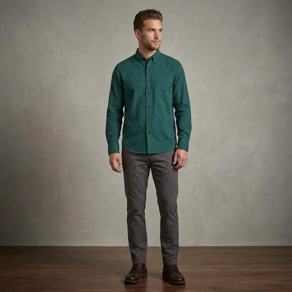 A man wearing a dark teal-green long-sleeve button-down shirt, gray trousers, and brown leather shoes, standing against a textured gray studio wall on a wooden floor.
