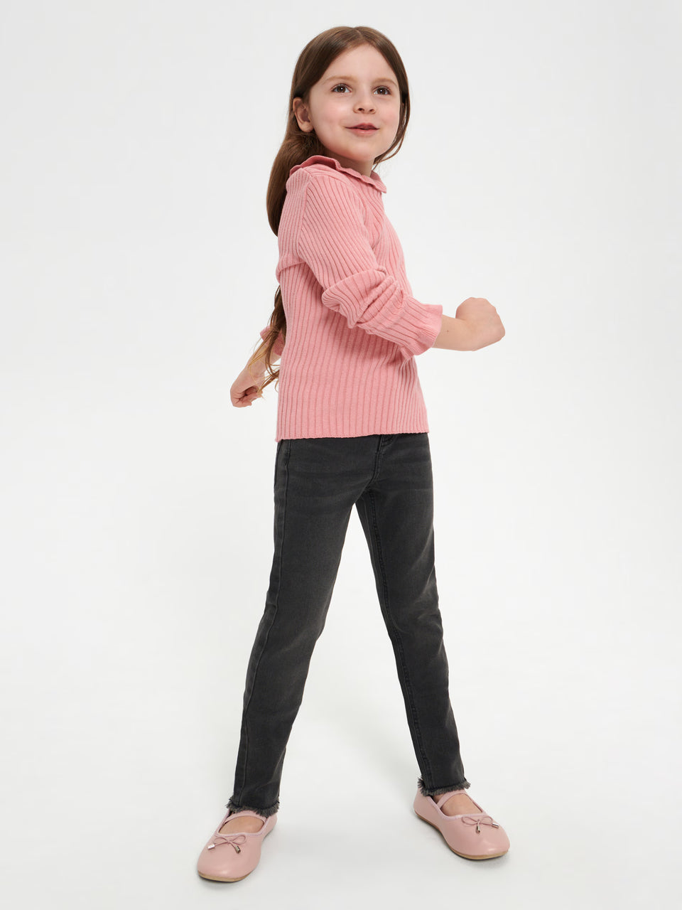 Young girl wearing a pink ribbed sweater and black pants on a white background