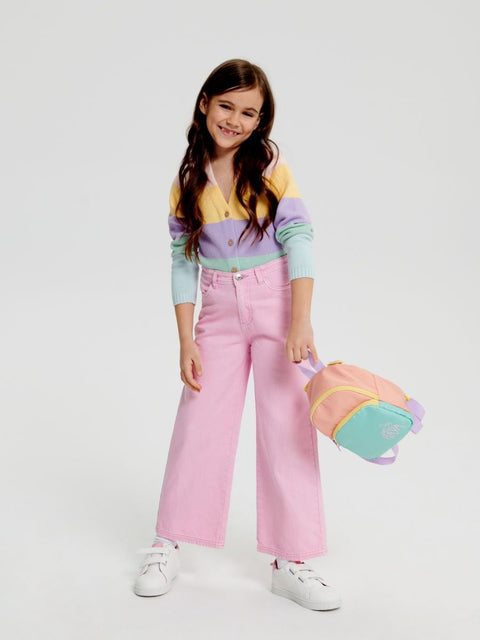 A girl wearing a multicolored top and pink wide-leg jeans, holding a multicolored toy and standing against a white background.