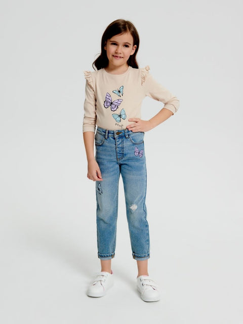 Young girl wearing a beige sweater with butterfly designs and blue jeans on a white background
