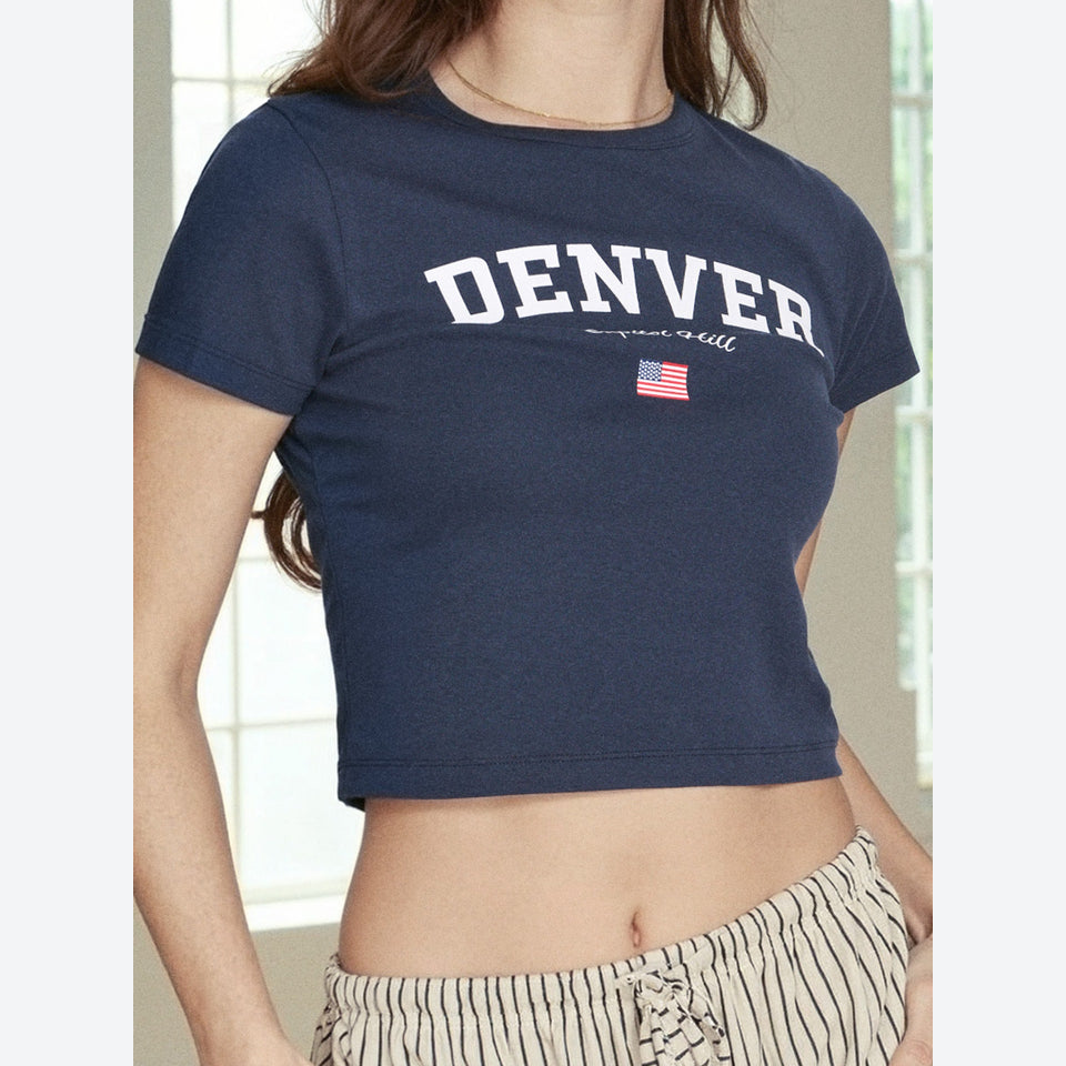 A woman wearing a navy blue cropped t-shirt with 'DENVER' printed in white arched letters across the chest, featuring a small American flag graphic below. The photo is cropped from the chest down, showing the model's midriff and striped pants against a softly blurred indoor background.