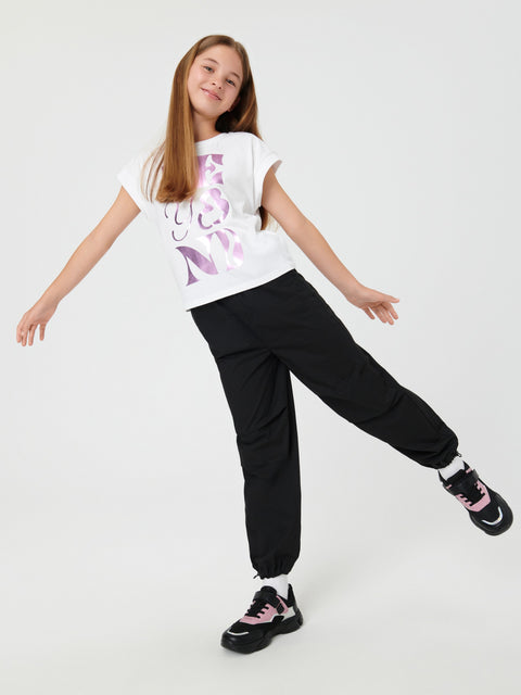 Young girl wearing a white t-shirt with a purple design, black pants, and black sneakers on a white background