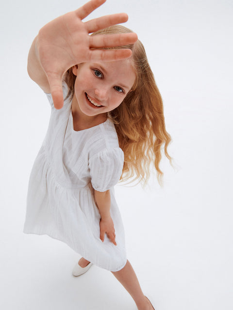 Young girl in a white dress posing with a hand over her face on a light background