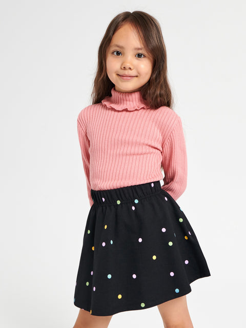 Young girl wearing a pink ribbed sweater and black skirt with colorful polka dots on a white background