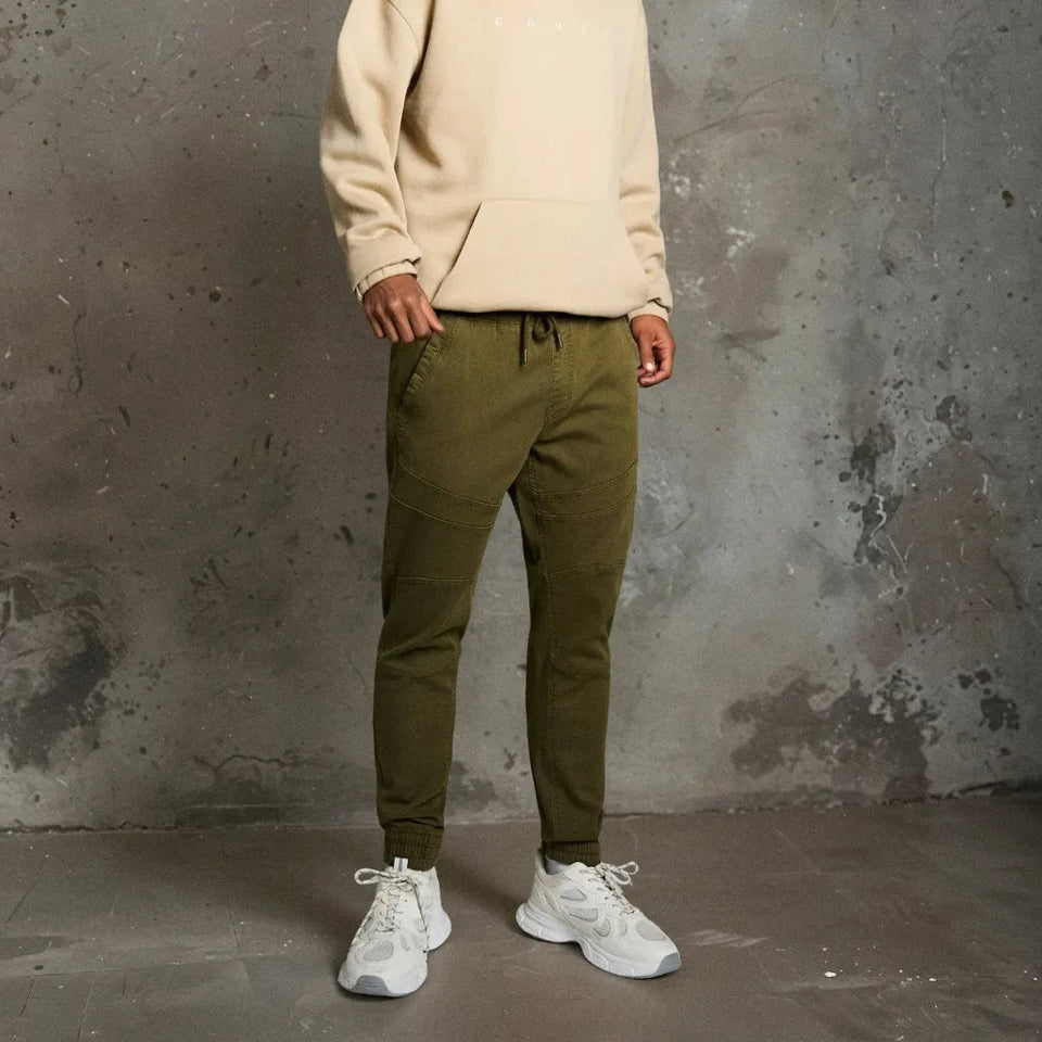 A person wearing olive green tapered jogger pants with a drawstring waist, styled with a cream hoodie and white sneakers against a textured gray concrete wall.