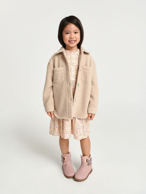 Young girl wearing a beige coat, floral dress, and pink boots on a white background