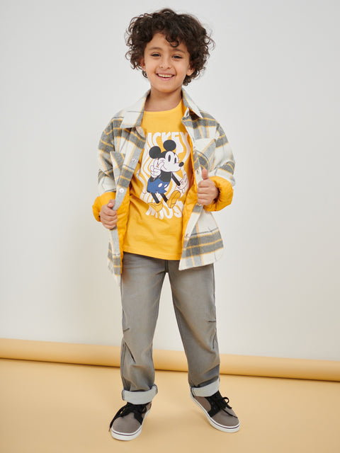 Child wearing a yellow shirt with a cartoon character, plaid jacket, and gray pants on a plain background