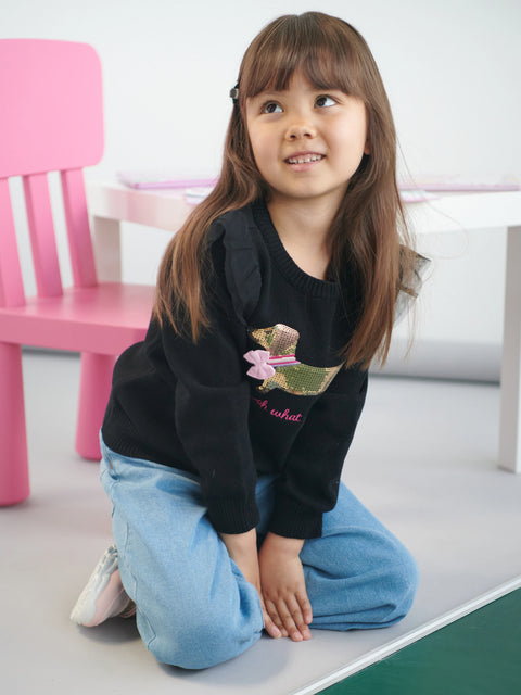 Young girl wearing a black sweatshirt with a teddy bear design, sitting on a white surface.