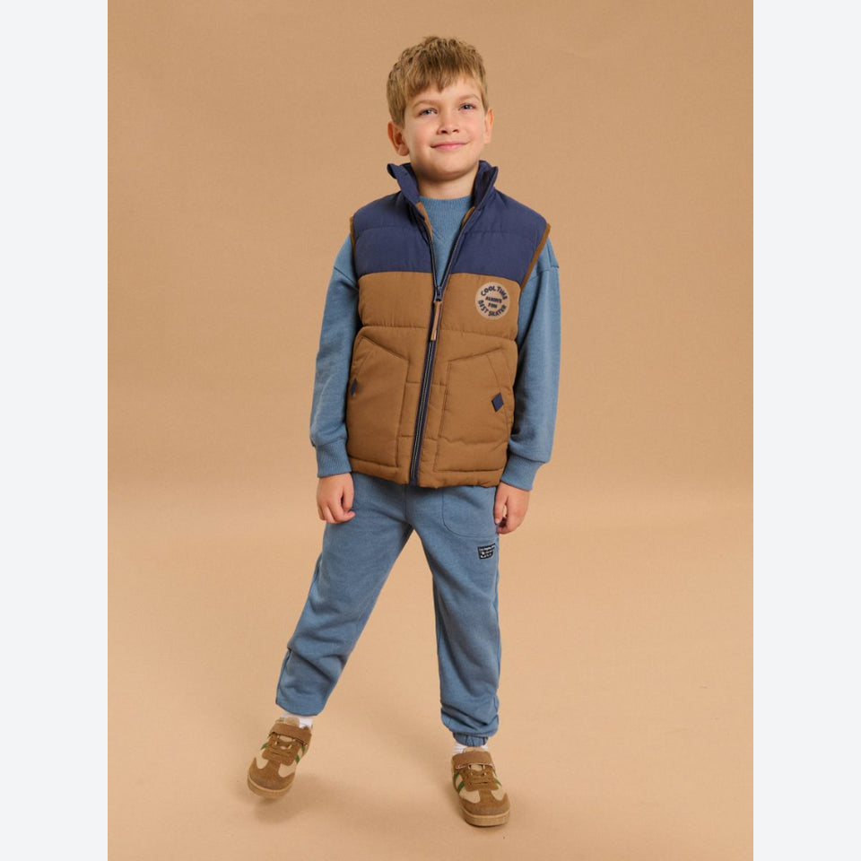 A young boy modeling a two-tone brown and navy quilted puffer vest over a blue long-sleeve shirt and matching blue sweatpants, standing against a solid tan studio background.