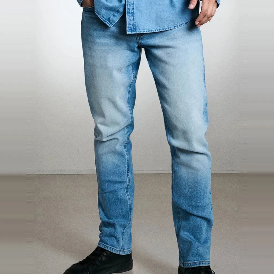 A person wearing light blue slim-fit denim jeans with a faded wash, paired with a matching light blue denim shirt and black shoes, shown from the waist down against a neutral studio background.