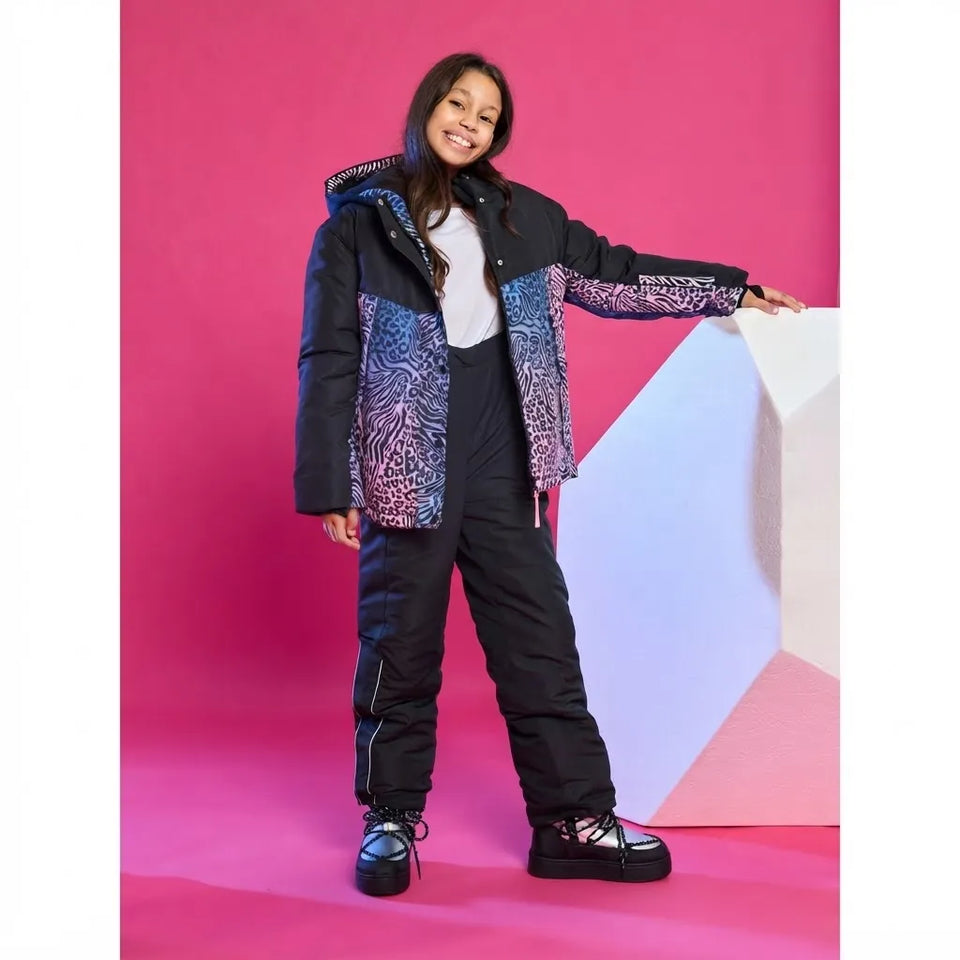 A child model wearing a black and purple patterned puffer jacket with matching black snow pants, posing against a vibrant pink studio backdrop with a white geometric prop.