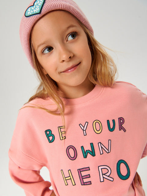 Young girl wearing a pink sweatshirt with 'Be Your Own Hero' text, looking to the side.