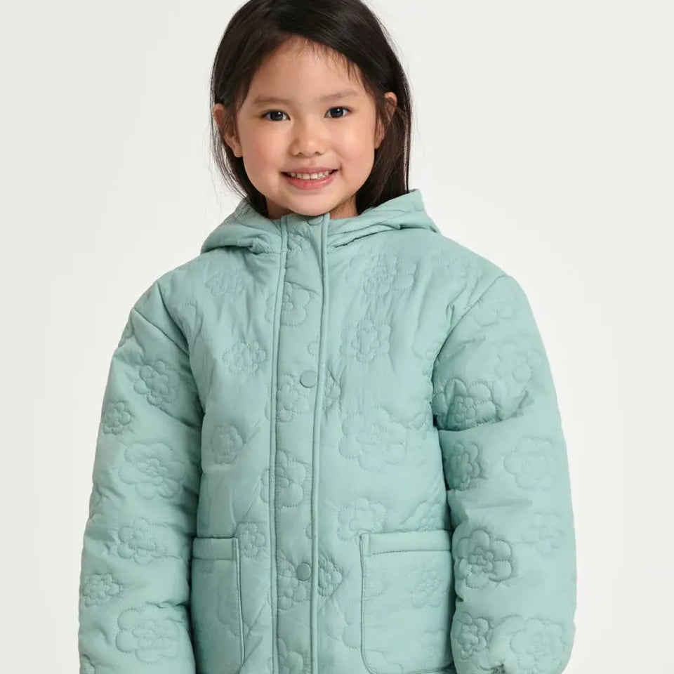 A young girl smiling while wearing a mint green quilted hooded jacket with a floral pattern, photographed against a plain light background.