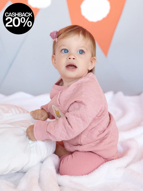 Baby in a pink outfit sitting on a soft surface with a colorful geometric background