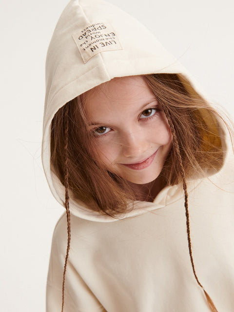 Young girl wearing a beige hooded garment with a visible brand label.