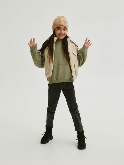 Person wearing a green hoodie, beige vest, black pants, and a beige beanie on a white background