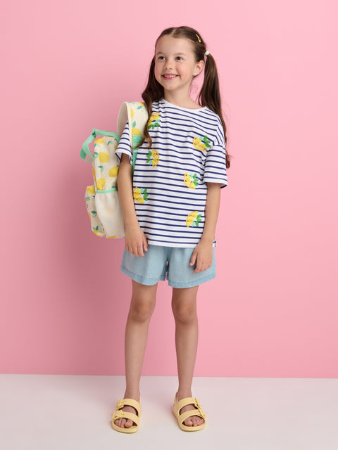 Young girl wearing a striped shirt with pineapple design, denim shorts, and yellow sandals, holding a colorful backpack against a pink background.