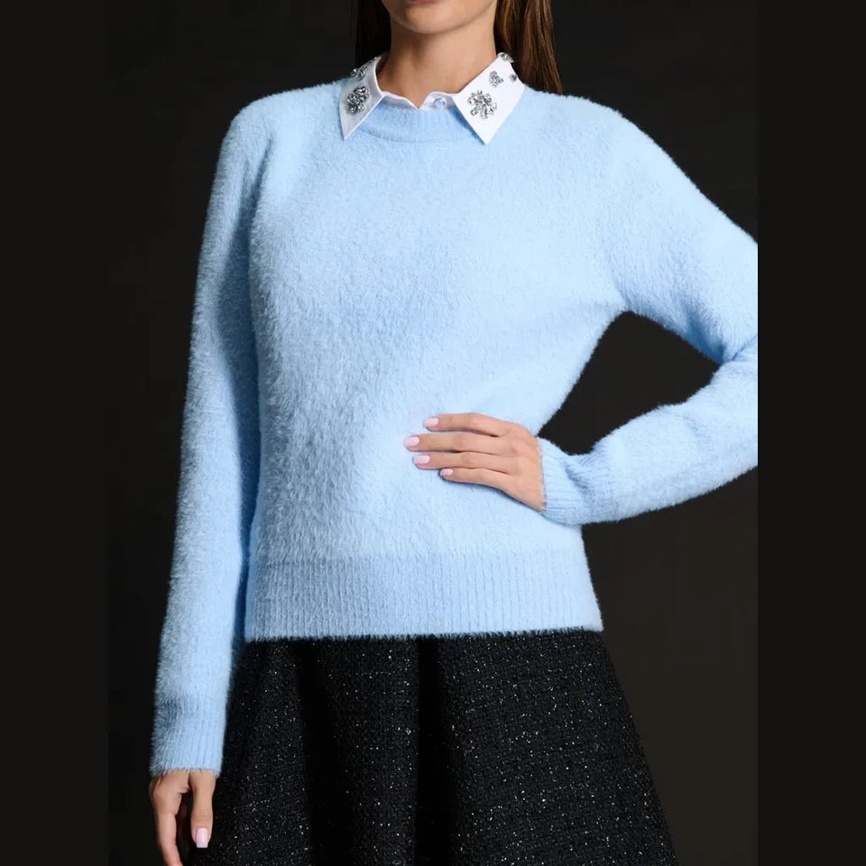 A woman wearing a light blue fuzzy knit sweater with a white decorative collar featuring black floral embroidery, paired with a black sparkly skirt, set against a dark studio background.