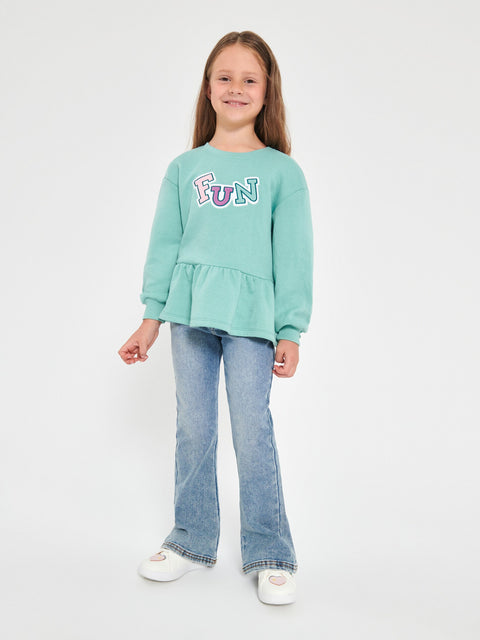 Young girl wearing a light blue sweatshirt with 'FUN' printed on it and blue jeans against a white background.