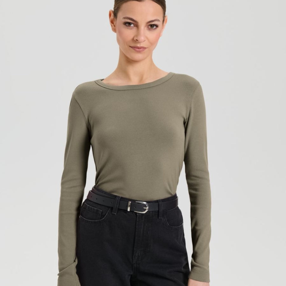 A woman modeling a long-sleeve crew-neck knit top in olive green, paired with black jeans and a black belt, against a light gray studio background.
