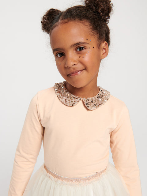 Young girl wearing a beige long-sleeve top with a decorative collar against a plain background