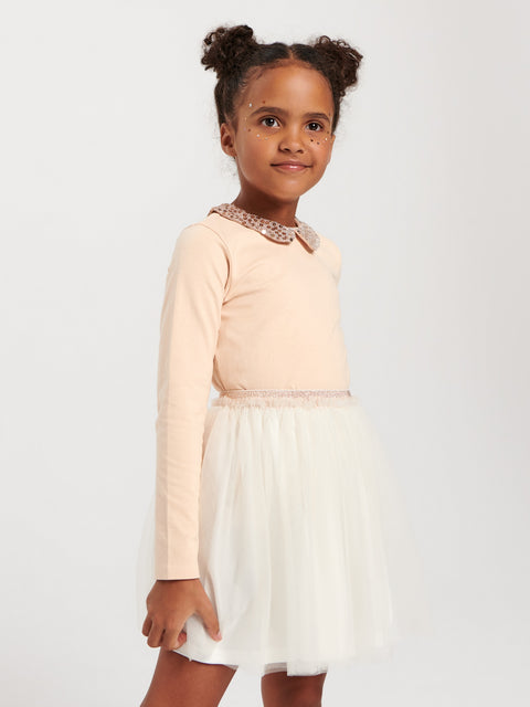 Young girl wearing a light pink long-sleeve top with lace collar and white skirt on a plain background