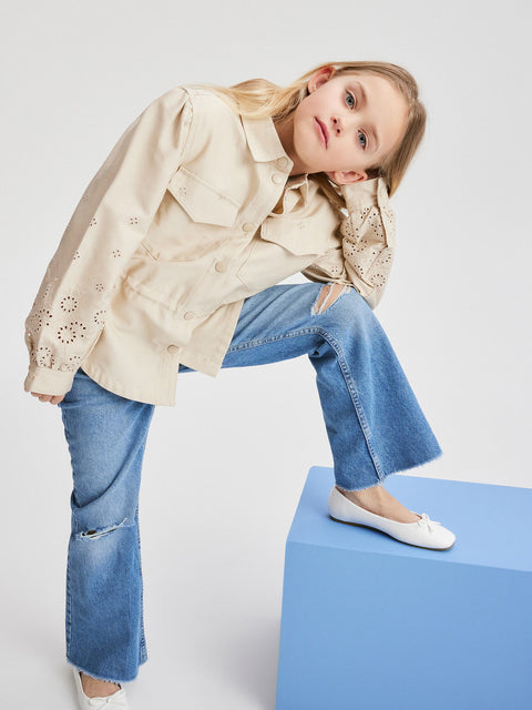 Young girl wearing a beige jacket and blue jeans sitting on a blue box against a white background