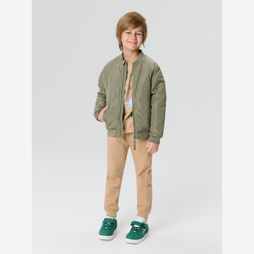 A young boy modeling an olive green bomber jacket over a tan graphic t-shirt, paired with tan pants and green sneakers, against a light gray studio background.