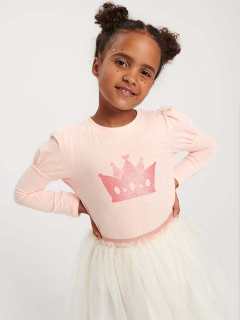 Young girl wearing a pink long-sleeve top with a crown design and a white skirt on a light gray background
