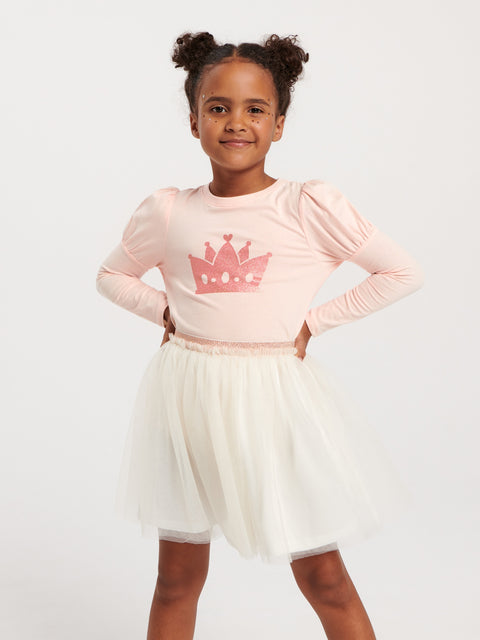 Young girl wearing a pink long-sleeve top with a crown design and a white skirt on a light gray background