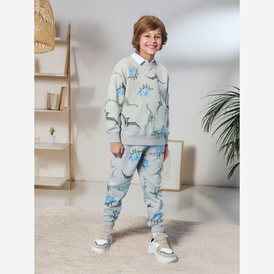 A child wearing a matching light blue sweatshirt and jogger pants set with a dinosaur print, styled with a white collared shirt underneath and white sneakers, standing in a bright, modern room.