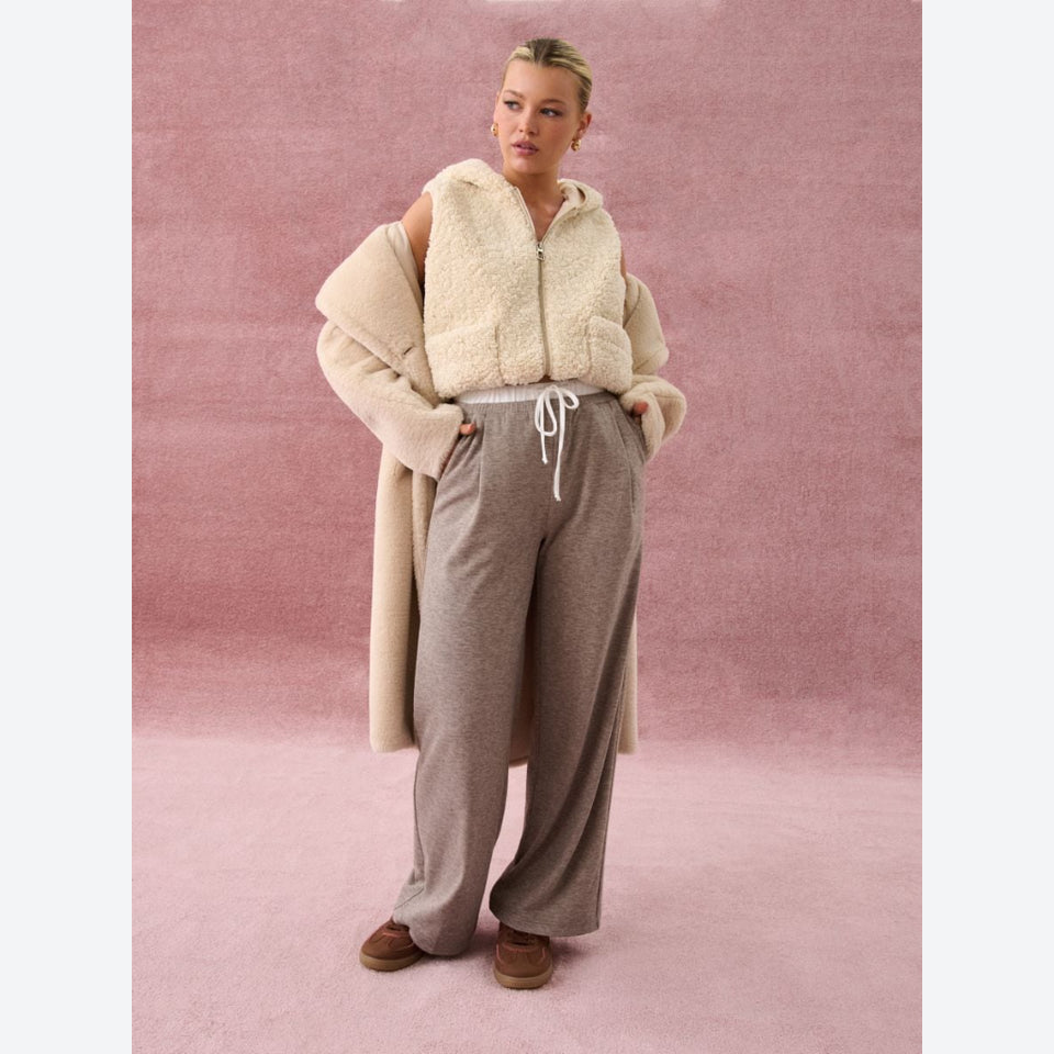 A woman models a cozy outfit consisting of a cream sherpa zip-up vest, a matching cream cardigan, heather taupe wide-leg sweatpants, and brown suede shoes, set against a textured dusty pink backdrop.
