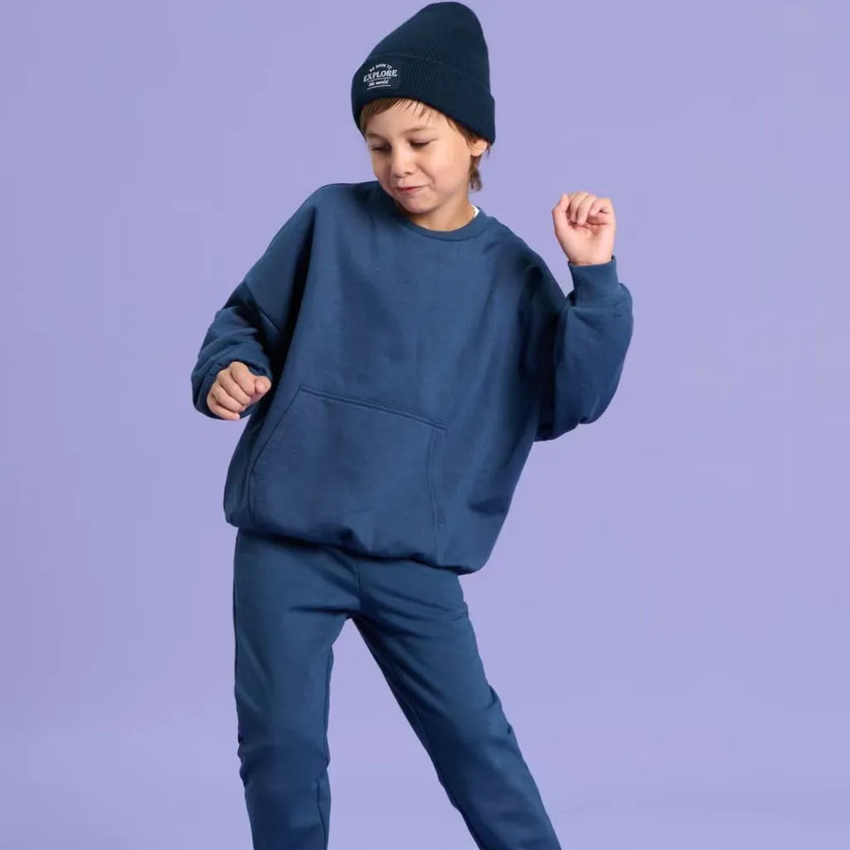 A child wearing a matching navy blue sweatshirt and jogger pants set, along with a navy beanie, posed against a solid lavender studio background.