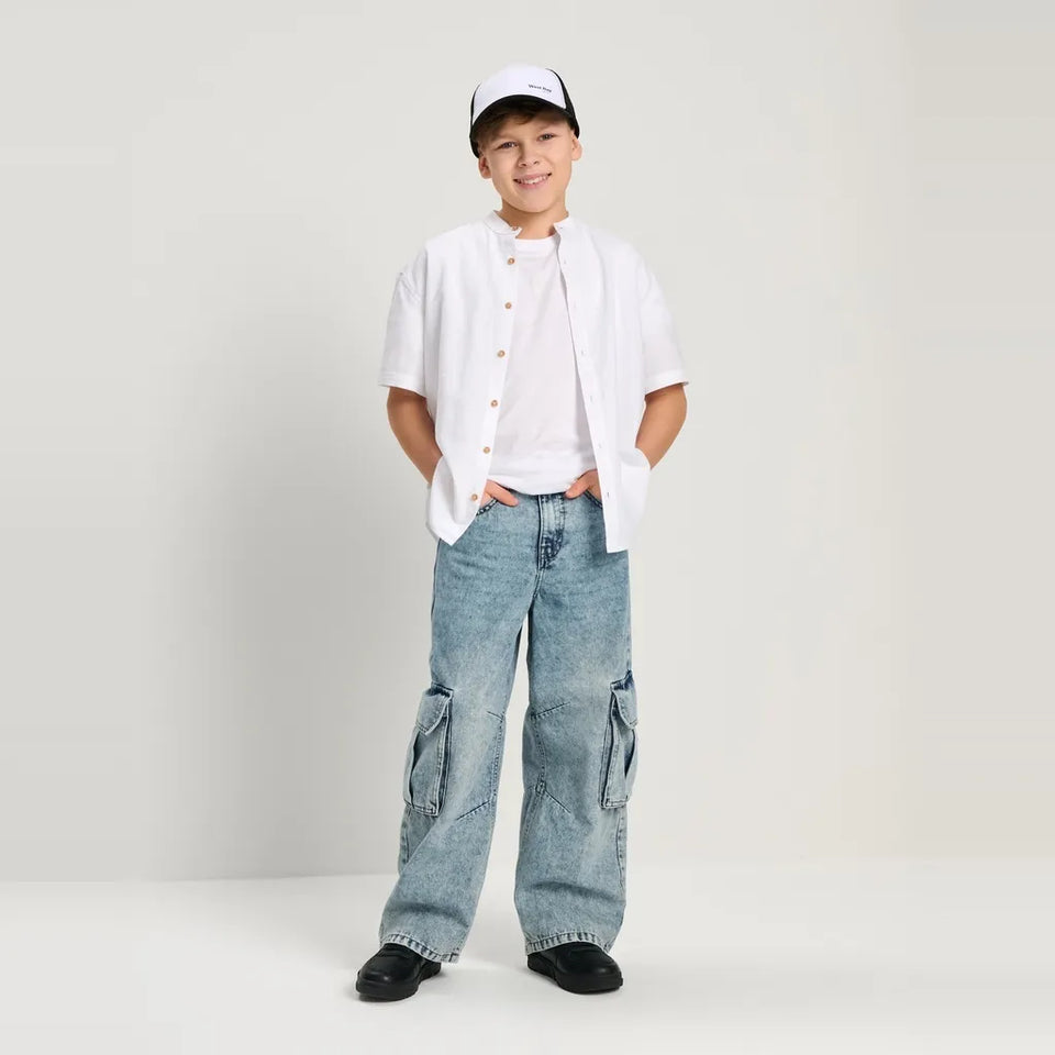 A young boy modeling a casual outfit consisting of a white short-sleeve button-up shirt, light-wash wide-leg denim cargo pants, a white t-shirt, black shoes, and a white and black baseball cap, standing against a plain light gray studio background.