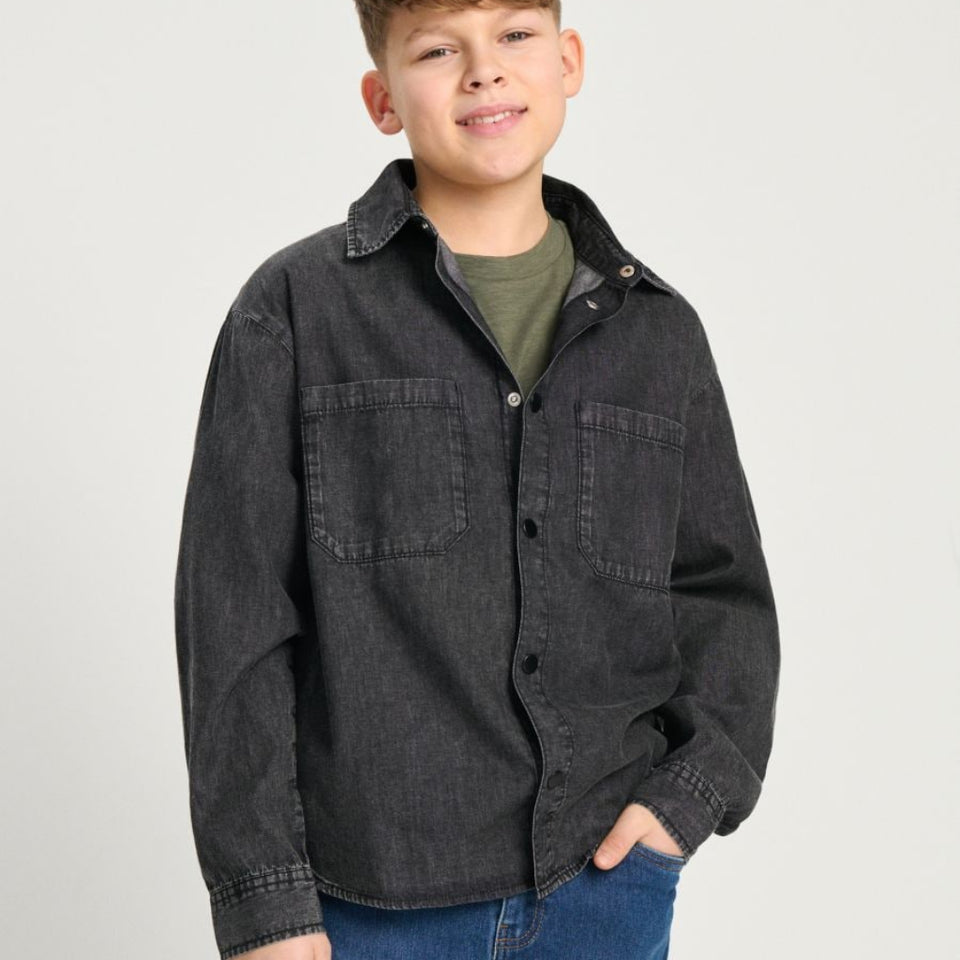 A boy modeling a dark charcoal gray button-up shirt jacket over an olive green t-shirt and blue jeans, photographed against a light gray studio background.