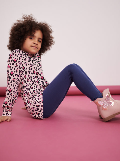 Child wearing a leopard print top and blue pants, sitting on a pink surface with a white background