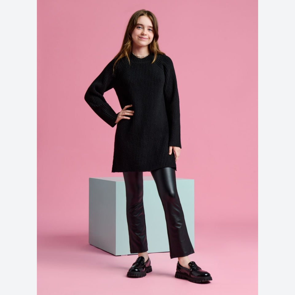 A young woman stands in a studio wearing a black ribbed long-sleeve tunic sweater, black flared pants, and black loafers, posed in front of a pink backdrop with a light blue-gray pedestal.