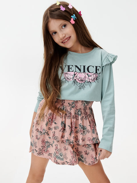 Young girl wearing a light blue sweatshirt with 'Venice' and floral designs, paired with a pink floral skirt on a white background.