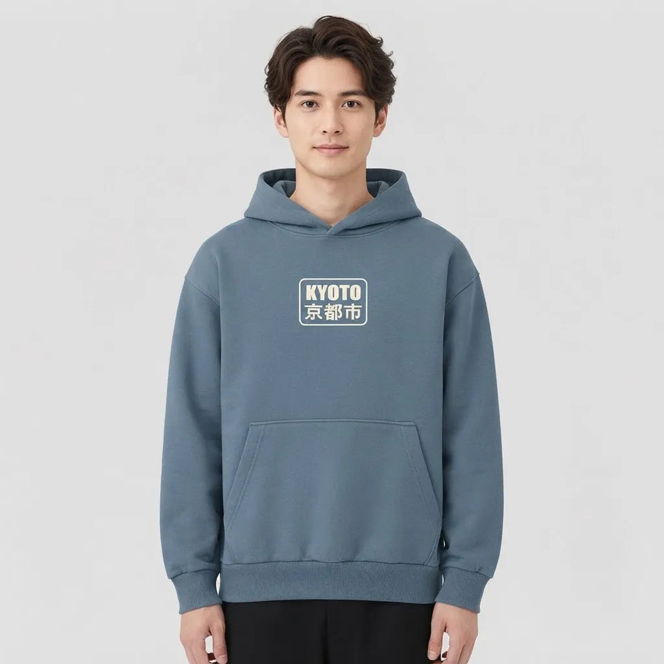 A man wearing a slate blue pullover hoodie with a white and black 'KYOTO 京都市' graphic on the chest, standing against a light gray studio background.