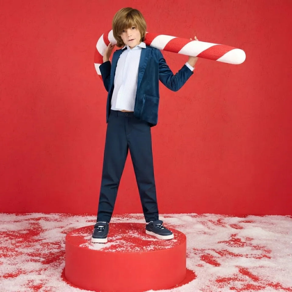 A child stands on a red pedestal against a red backdrop, wearing a navy blazer, white shirt, navy trousers, and dark sneakers, while holding a large red-and-white candy cane. White faux-snow covers the floor.