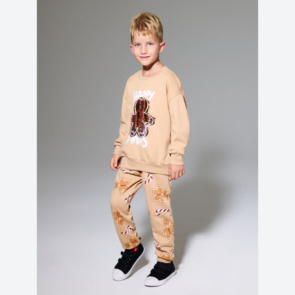 A young boy modeling a beige holiday-themed sweatshirt and jogger pants set. The sweatshirt features a gingerbread man graphic with 'MERRY CHRIST' in white text, and the joggers are decorated with gingerbread men and candy cane patterns.