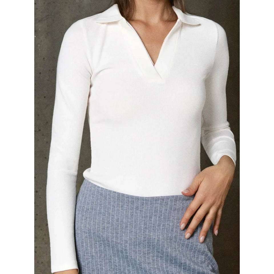 A woman wearing a fitted white long-sleeve polo-style top with a split V-neckline, paired with a blue textured skirt, shown against a dark gray wall.