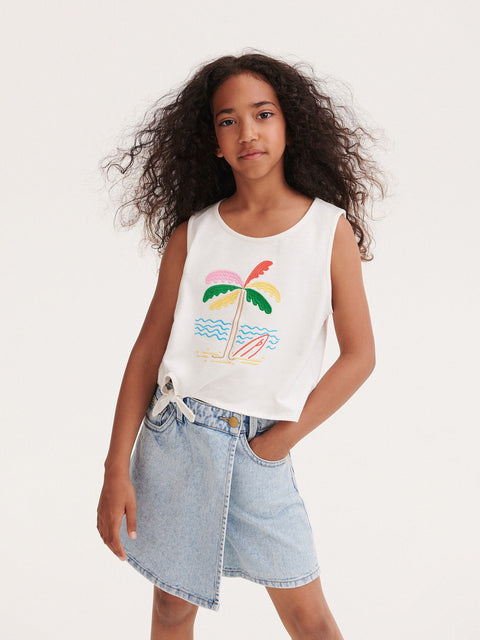 Young girl wearing a white tank top with a palm tree design and a denim skirt on a plain background