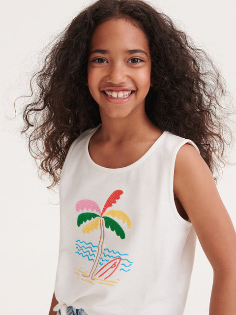 Young girl wearing a white sleeveless top with a colorful palm tree design on a plain background