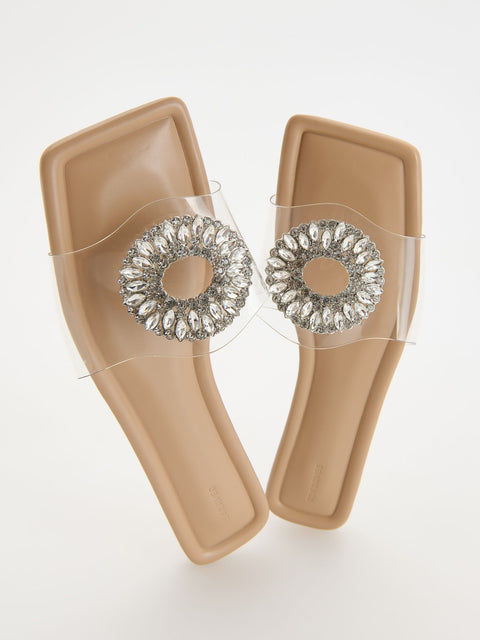 Pair of beige sandals with clear straps featuring decorative rhinestone embellishments on a white background.
