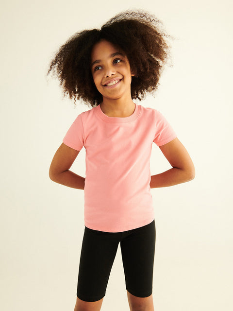 Young girl wearing a pink t-shirt and black shorts against a plain background